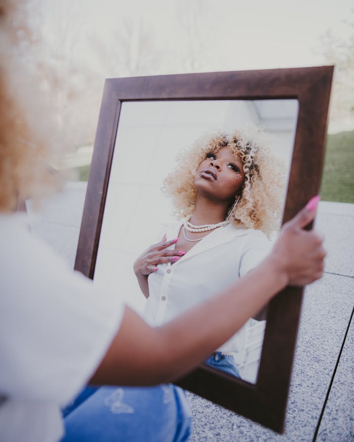 Offerings A fashionable woman with blonde curly hair holding a mirror outdoors, reflecting her confident look.